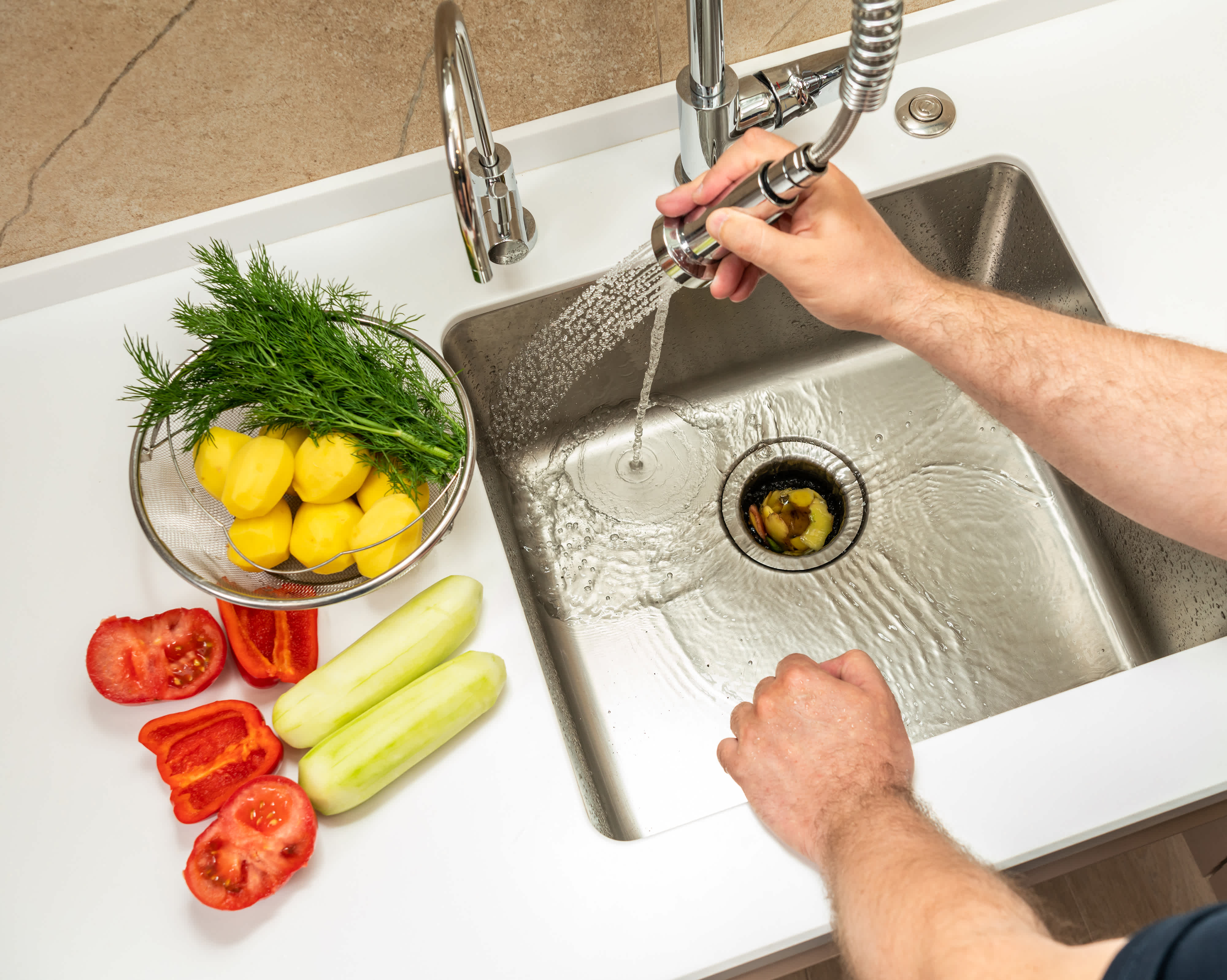 Person rinsing a kitchen sink with a pull-out faucet while peeled potatoes, cucumbers, red peppers, and fresh dill sit on the counter