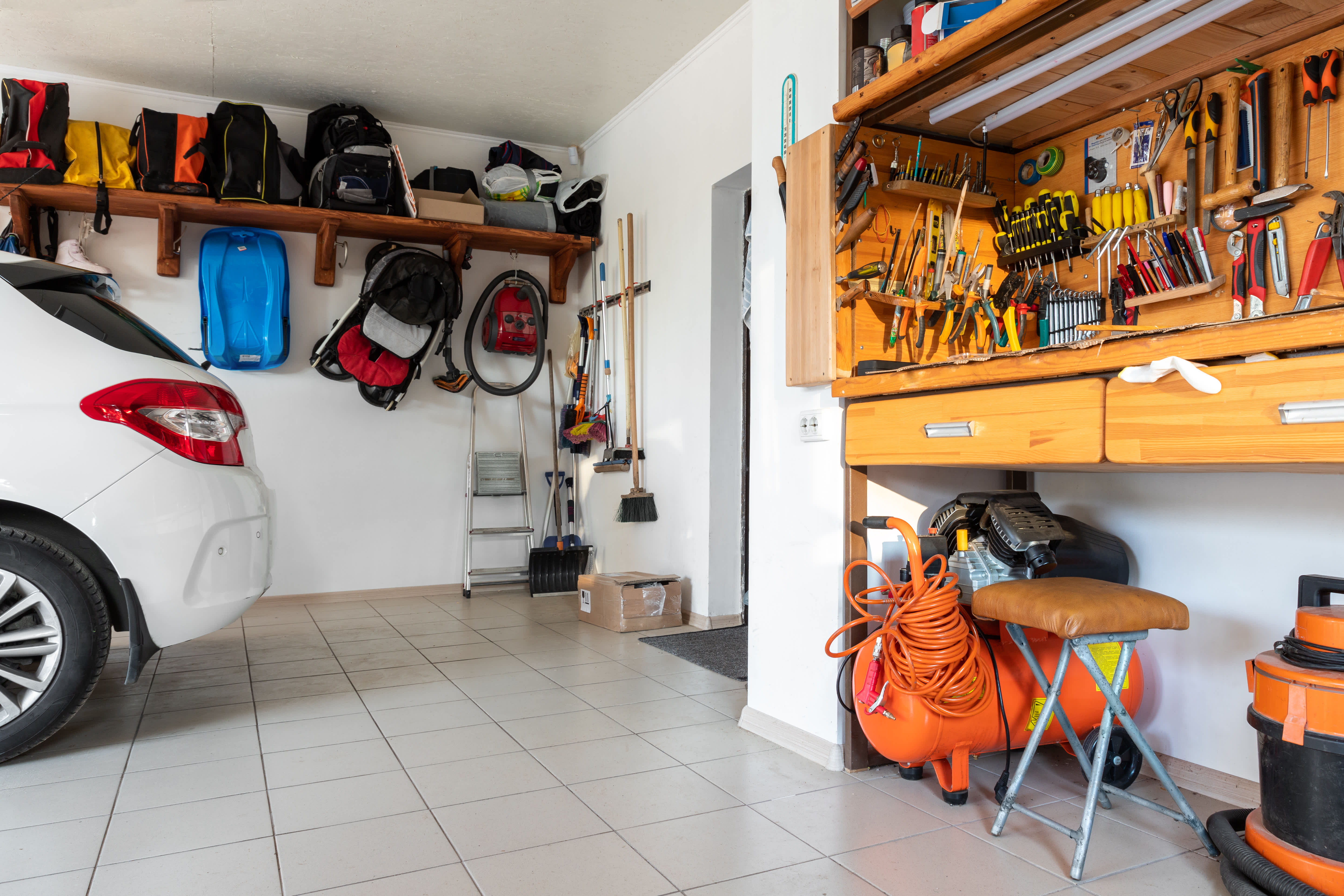 Organized garage with a white car, wall-mounted shelves holding bags, a workbench filled with tools, and cleaning equipment neatly arranged