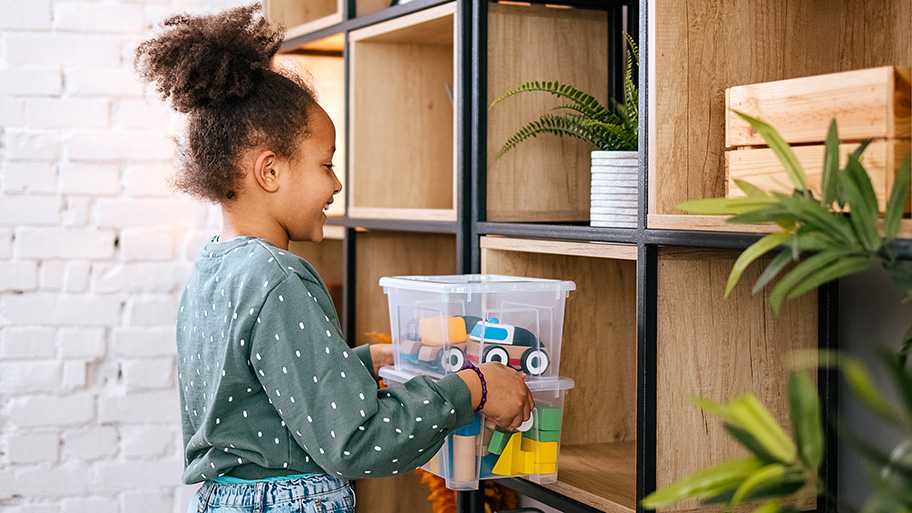 Smiling young girl organizing toys in a plastic storage bin while putting it on a wooden shelf at home