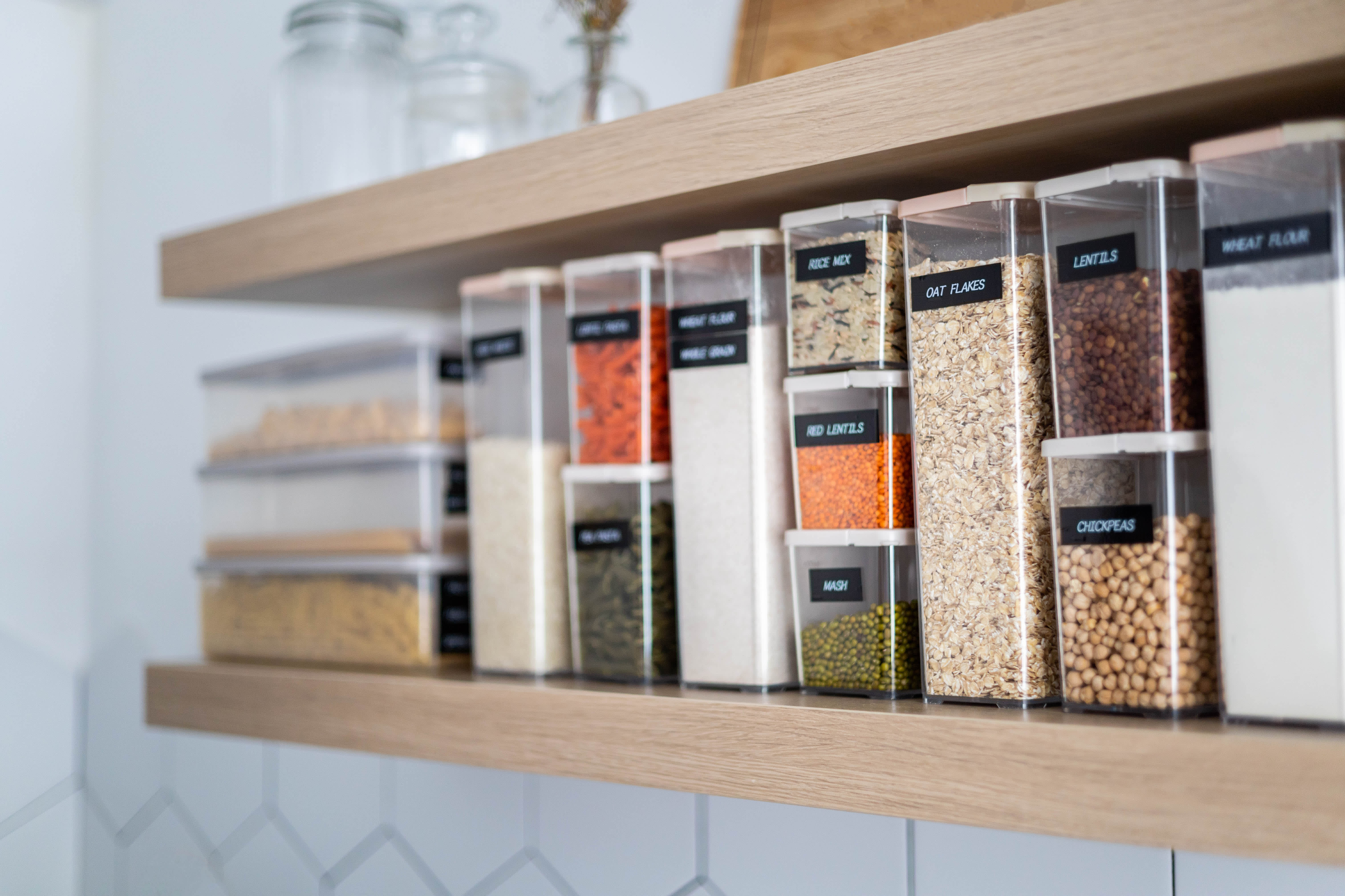 Close-up of labeled plastic containers filled with dry food items such as pasta, rice, lentils, oats, and chickpeas neatly arranged on a wooden kitchen shelf