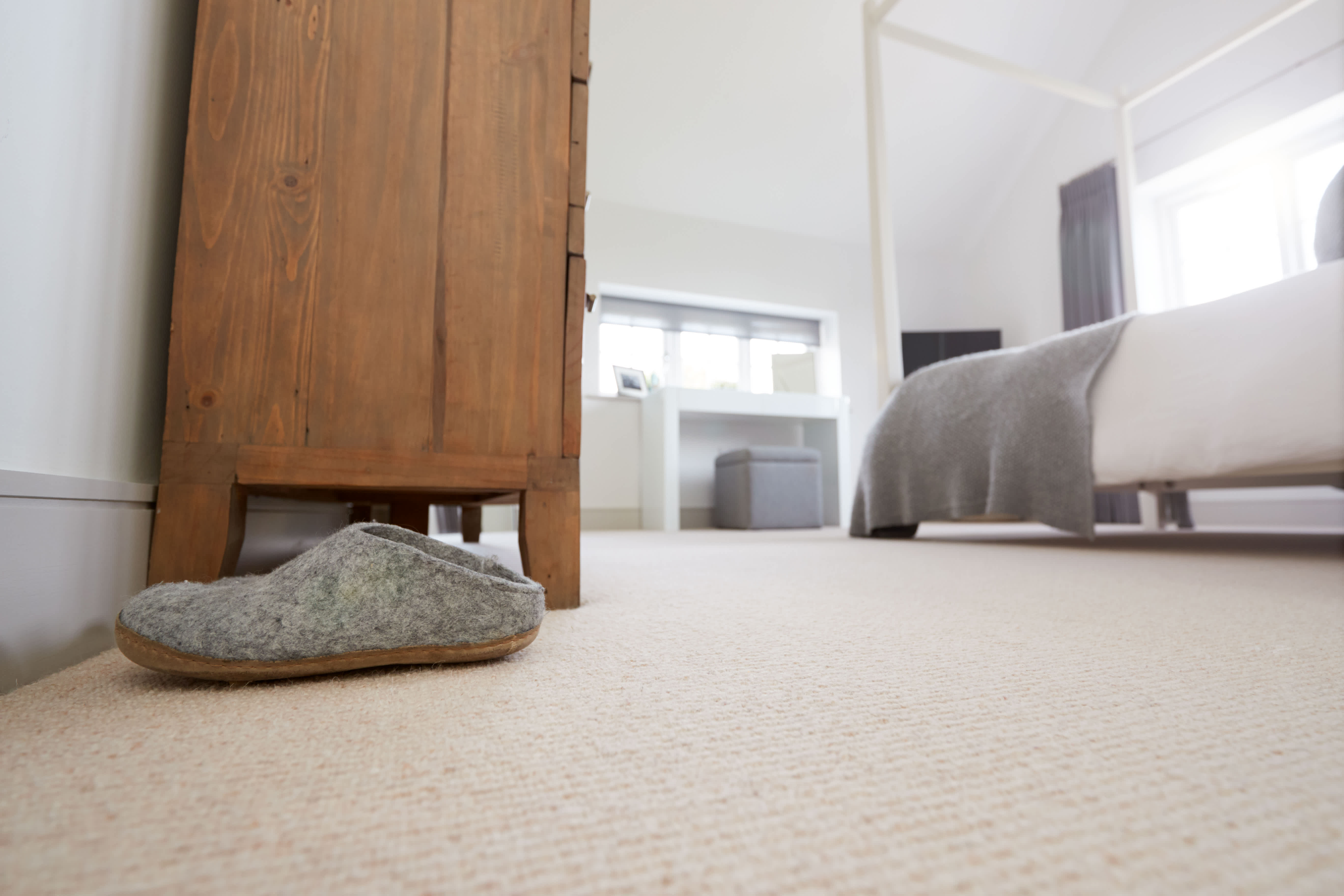Cozy bedroom with a single gray slipper on a soft carpet near a wooden dresser, with a neatly made bed and natural light from windows in the background