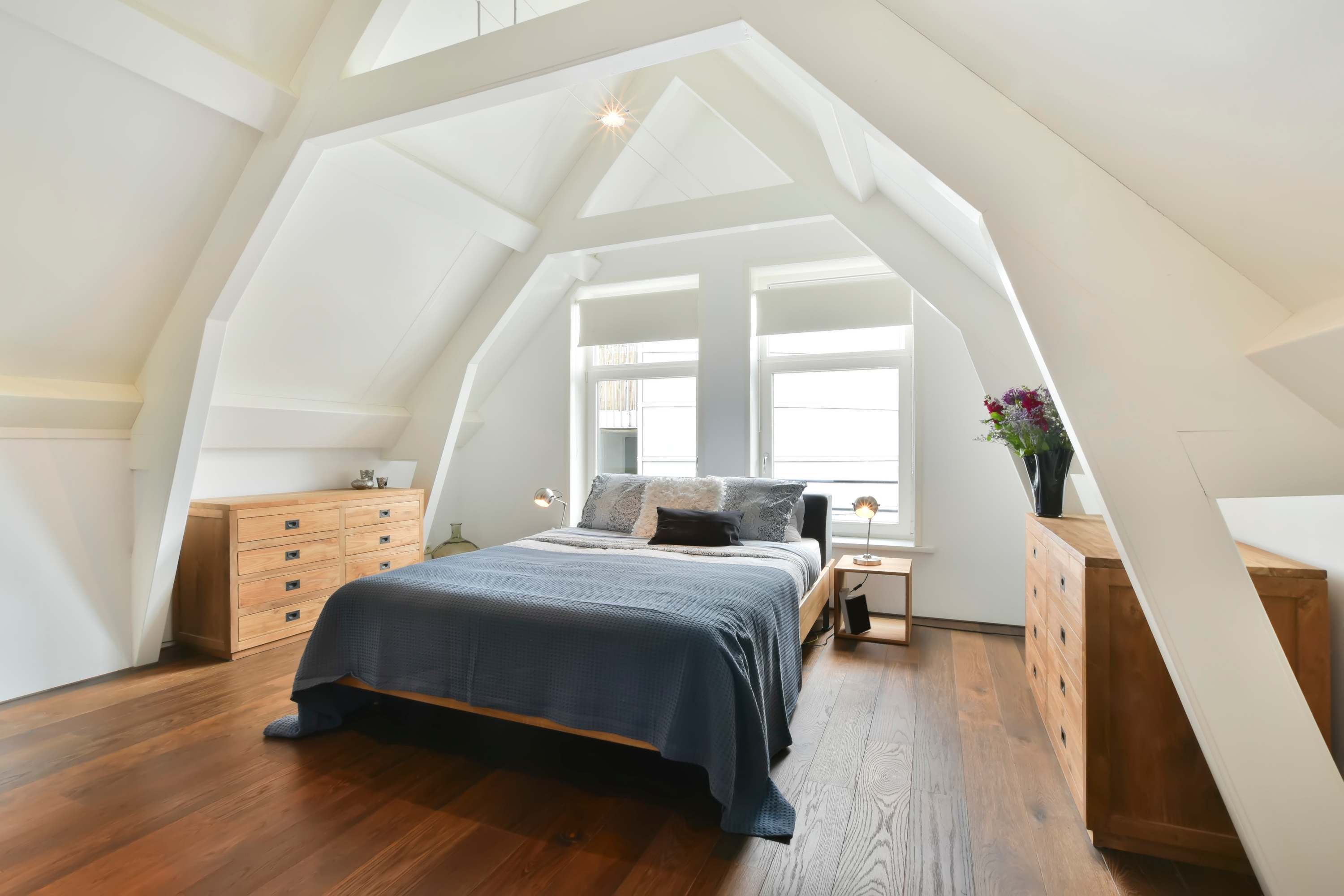Bright attic bedroom with sloped ceilings, wooden floors, a double bed with gray bedding, and wooden dressers on each side