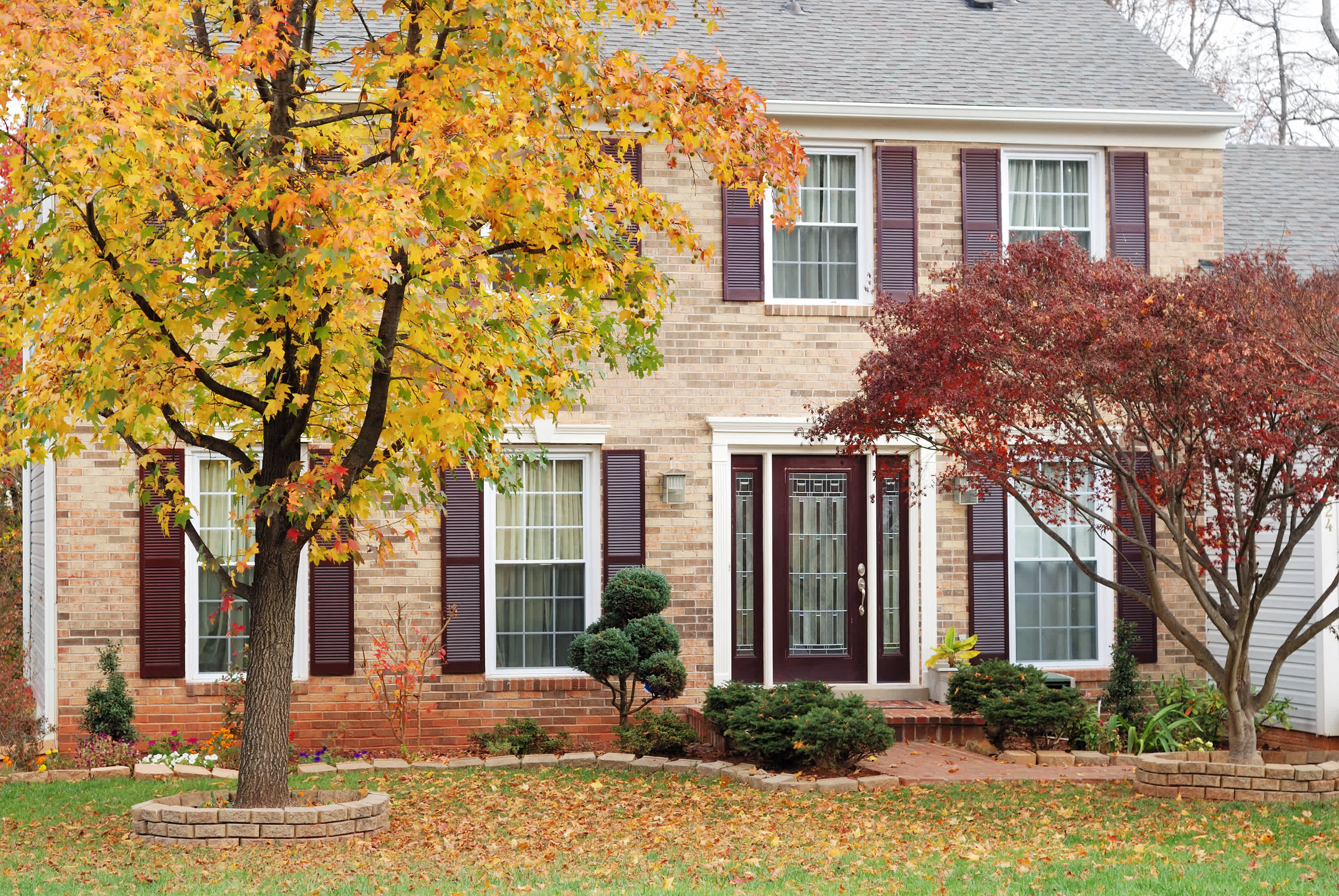 Traditional brick house with white trim, dark shutters, and a landscaped yard with autumn trees
