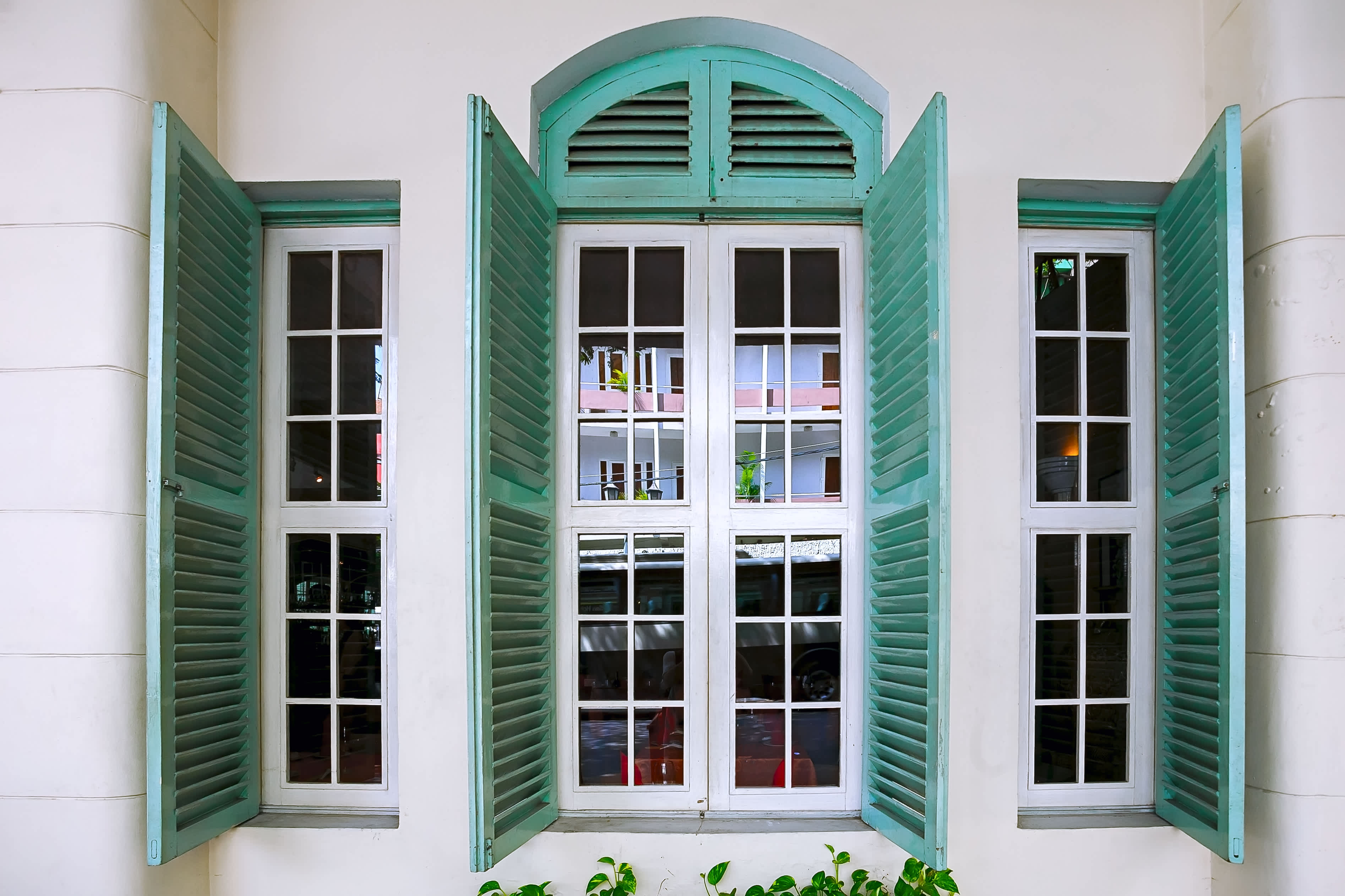 White wall with tall windows framed by teal wooden shutters, partially open to reveal reflections of nearby buildings