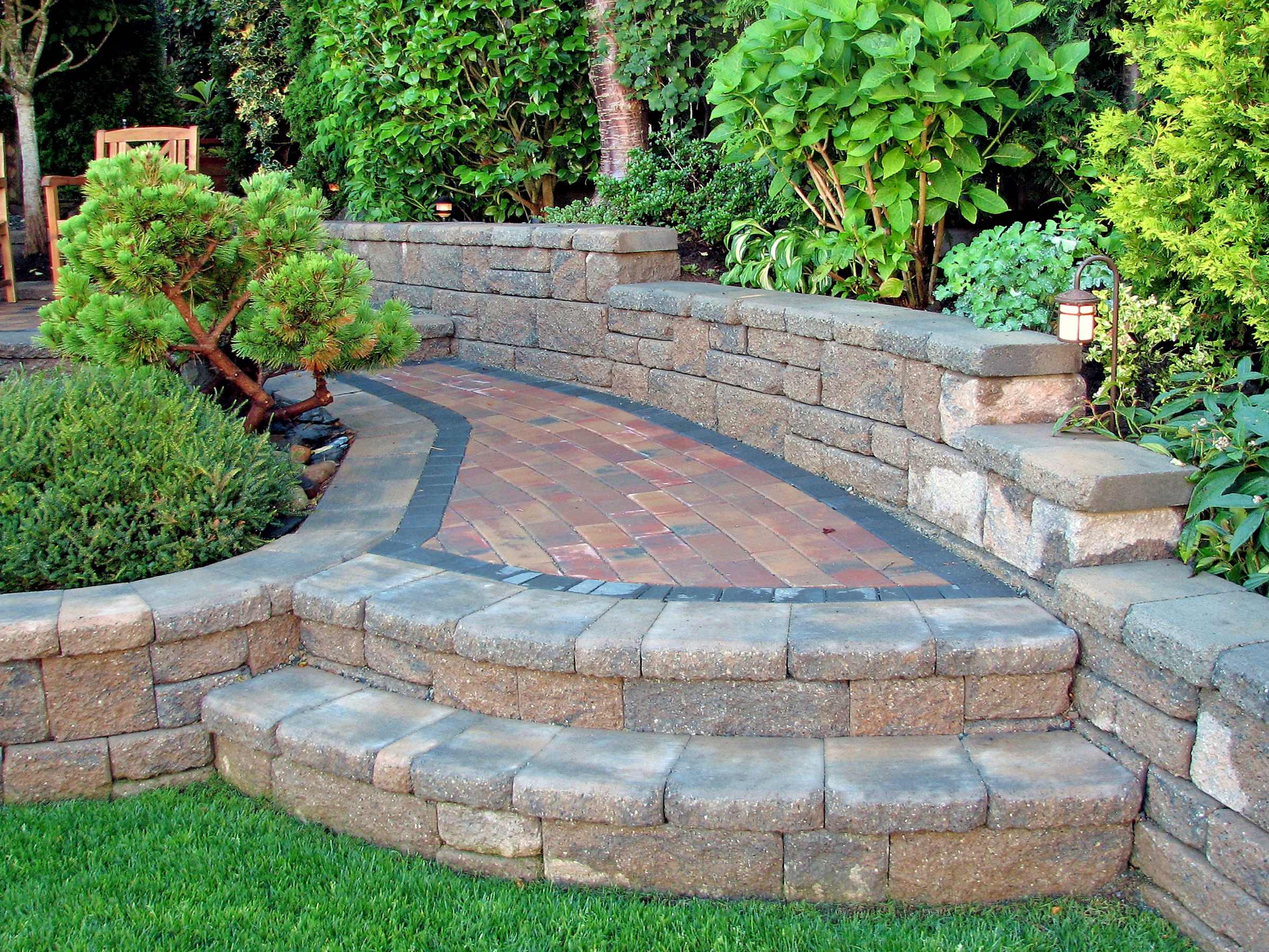 Curved stone retaining wall with built-in steps leading to a brick pathway surrounded by lush greenery