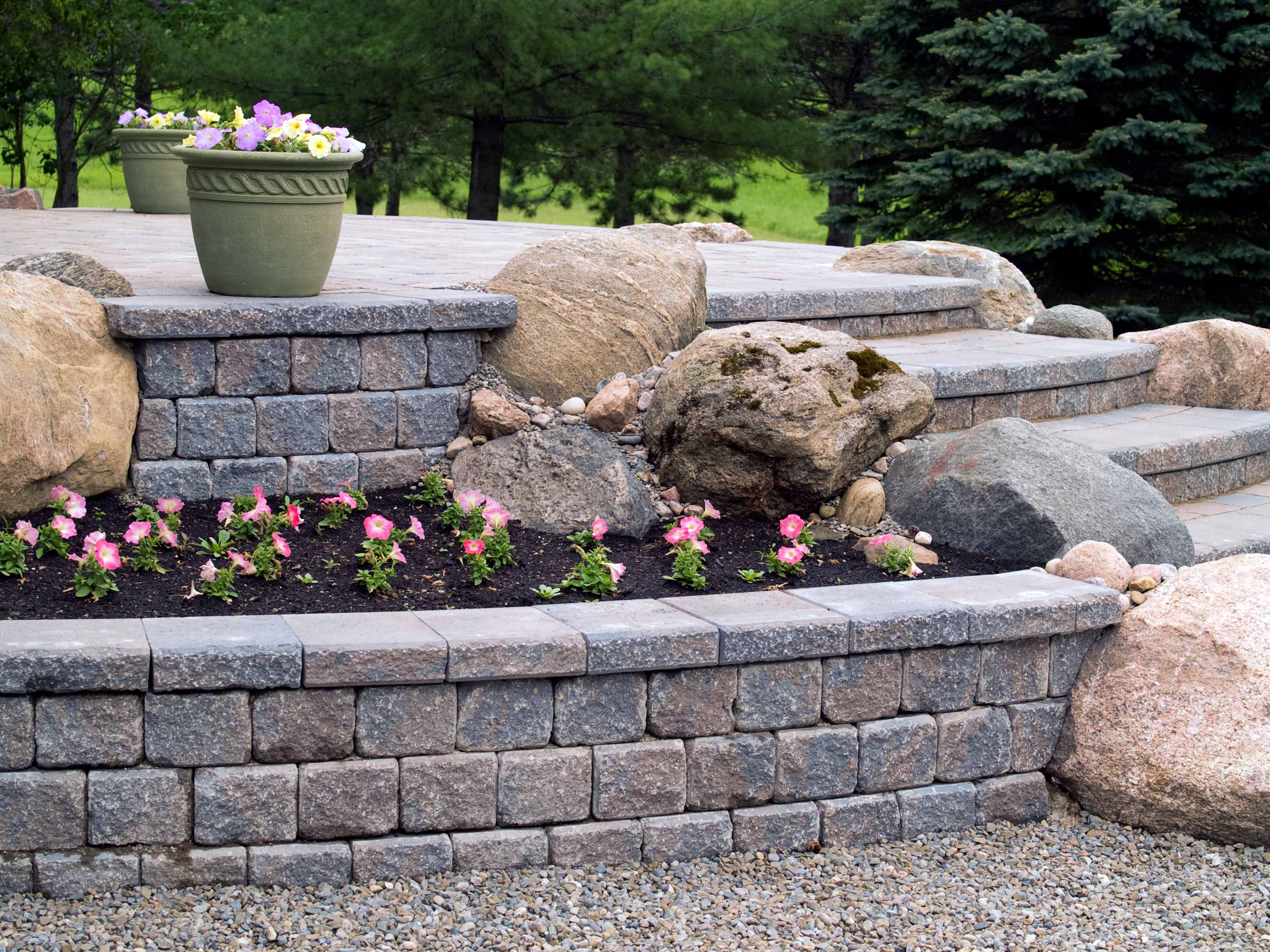 Stone retaining wall with built-in steps, accented by large boulders, potted plants, and blooming pink flowers