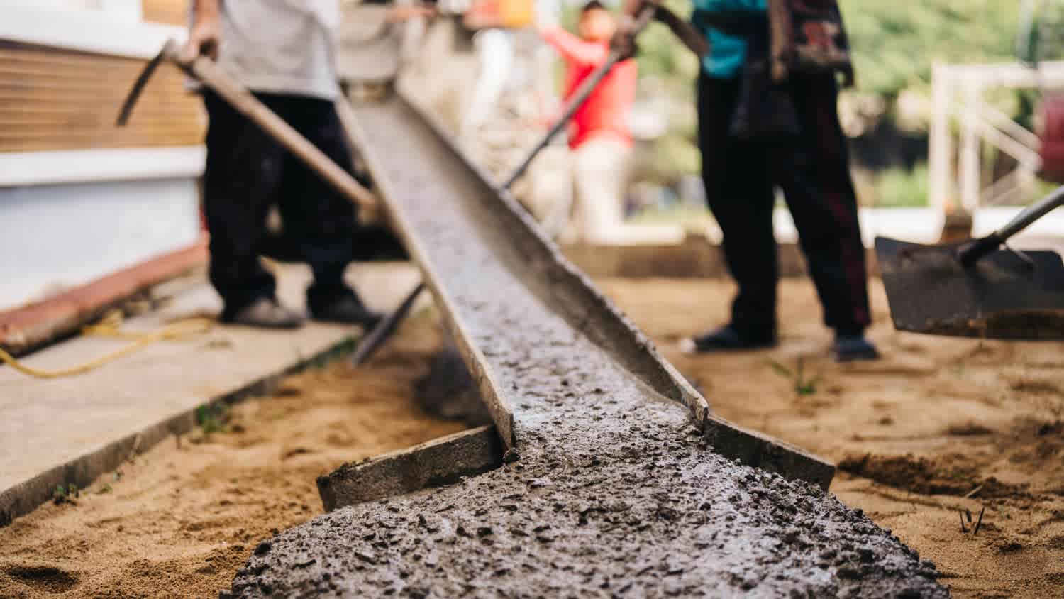 Wet concrete flows from a chute onto the ground as workers spread and level it at a construction site, preparing a foundation