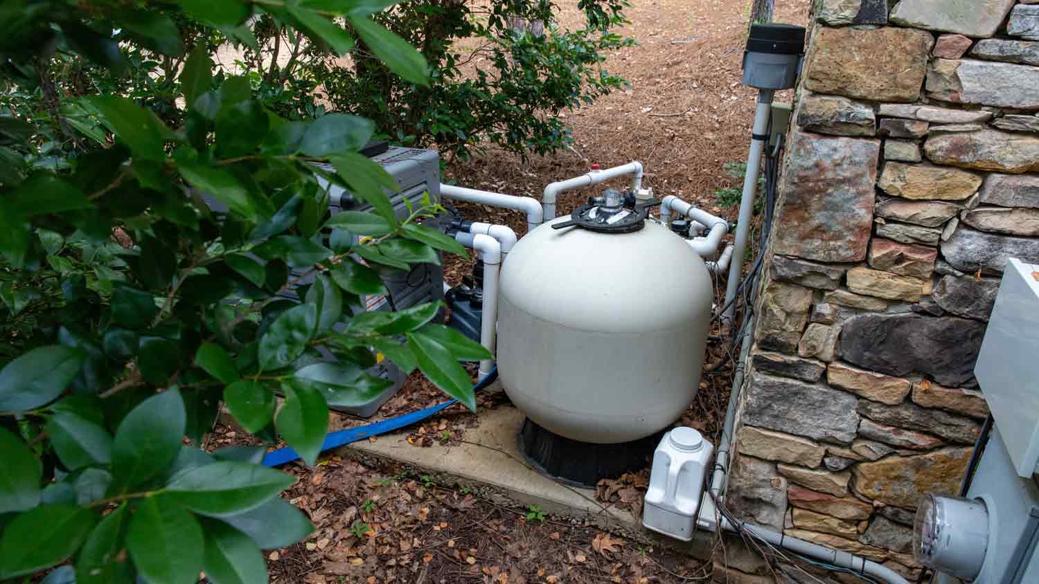 A residential pool pump on the ground outside next to a bush and a stone wall.