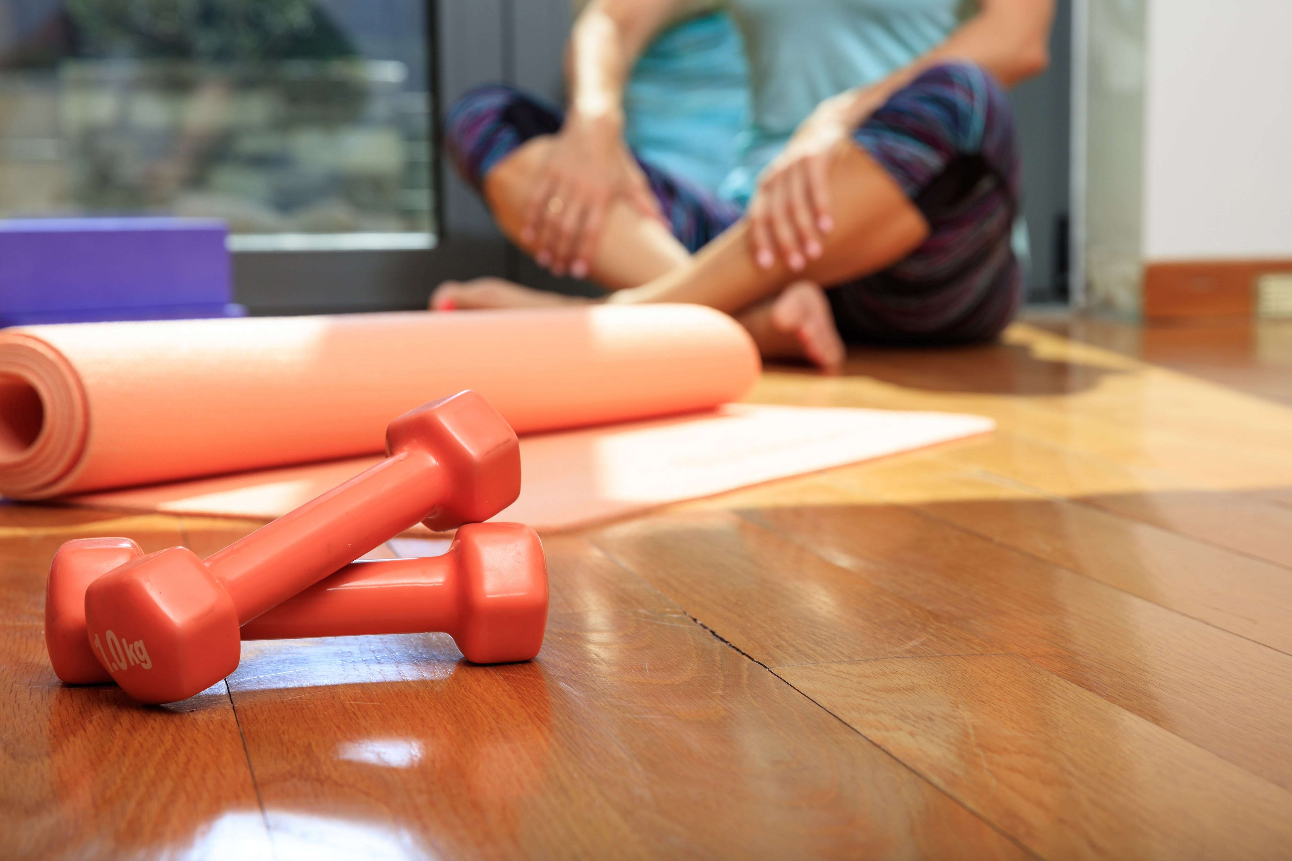 A woman in workout clothes sits on a wooden floor next to an orange yoga mat and dumbells