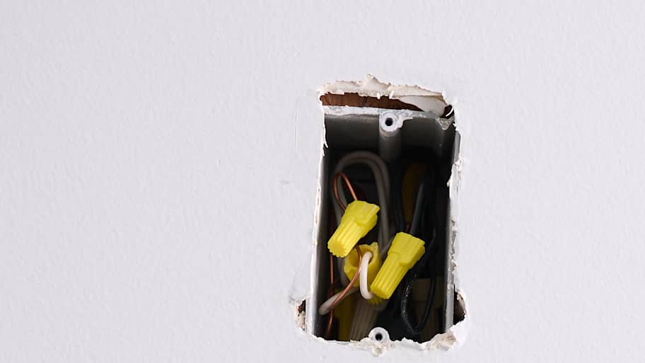 An open electrical outlet with exposed wires is set in a white wall, with drywall dust on the floor, showing ongoing electrical installation work.