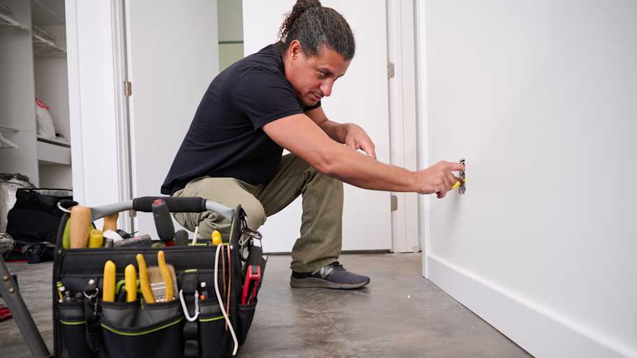 A technician kneels on the floor while working on an electrical outlet installation, with a tool bag full of equipment nearby for home repairs.