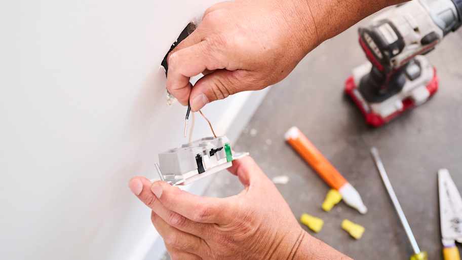 Close-up of a technician's hands connecting wires to a wall socket, with tools, a power drill, and wiring supplies on the floor nearby.
