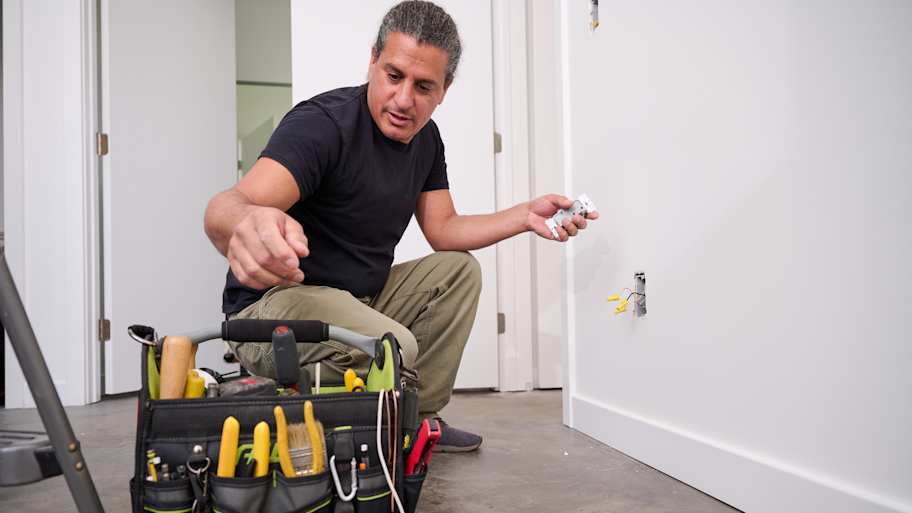 A technician kneels to install an electrical outlet, holding the outlet in one hand while selecting tools from a nearby tool bag for the job.