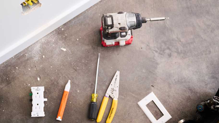 A set of tools, including a power drill, screwdriver, wire stripper, and electrical socket, are neatly arranged on a concrete floor beside a wall opening.