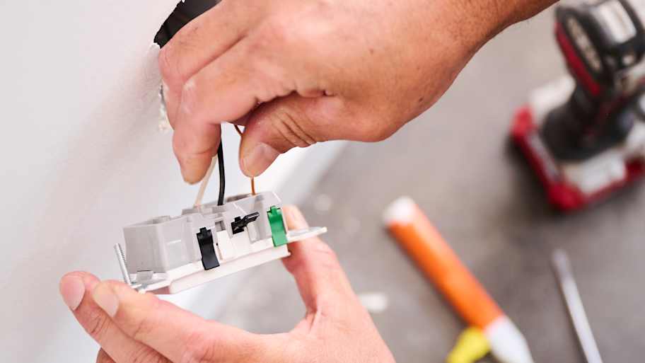Close-up of a technician's hands connecting black and copper wires to a wall socket, with a power drill and other tools in the background.