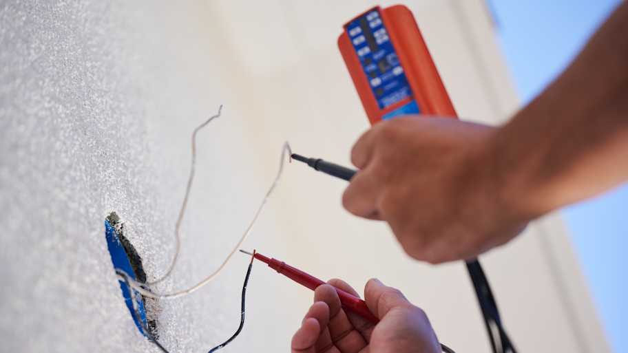 A close-up of a technician testing exposed wires with a multimeter to ensure proper electrical connectivity on a white wall installation.