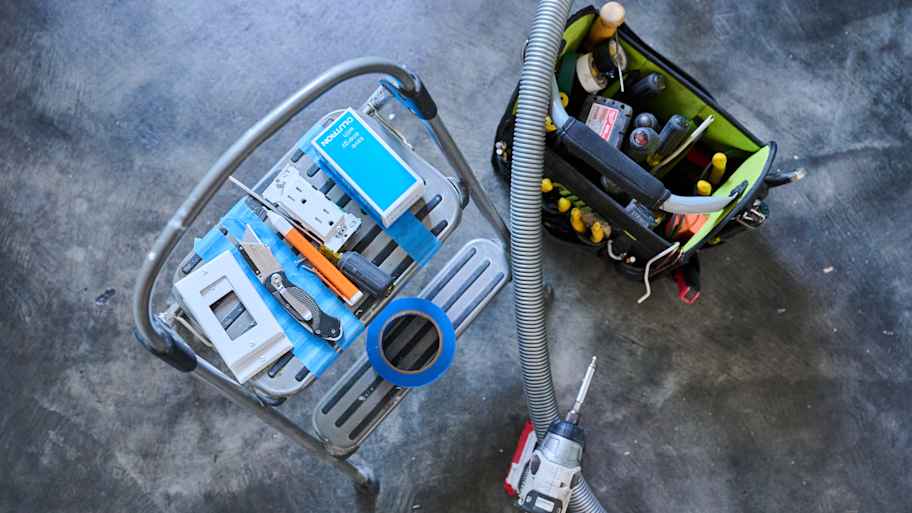 A step stool holds electrical tools, outlet covers, and tape, while a tool bag and drill sit nearby, indicating electrical work in progress.