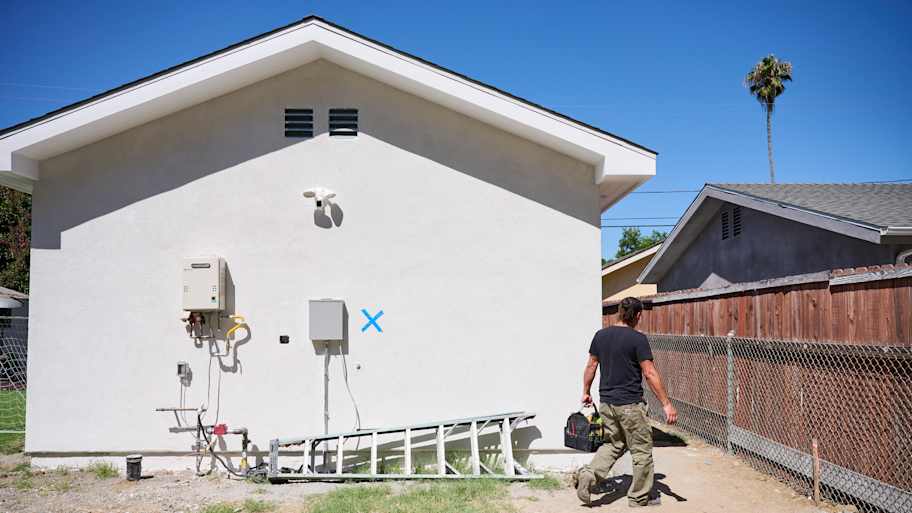 A technician in a black shirt and khaki pants walks away from a house wall with utility boxes, a ladder, and a blue "X" marking on it.