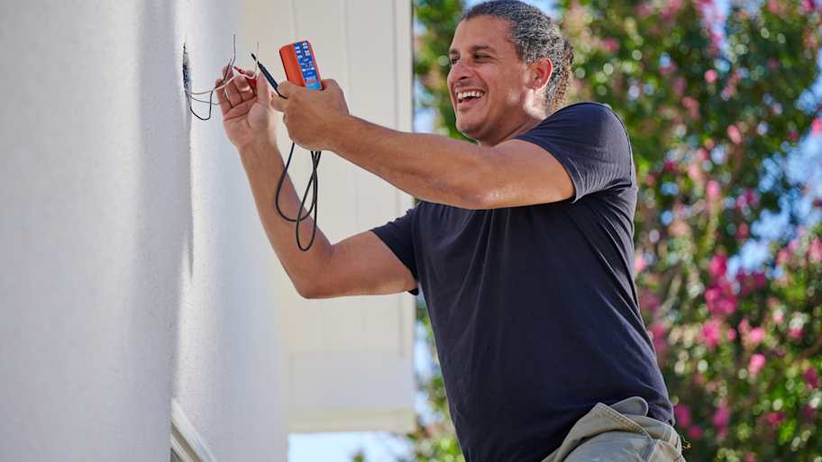 Technician smiling while using a multimeter to test wiring on an outdoor wall, enjoying his work on a sunny day with a lush background.