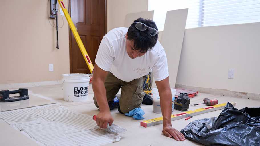 A worker applies tile adhesive to the floor with a trowel, preparing the surface for tile installation in a home renovation project.