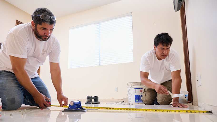 Two workers measure and prepare the floor for tile installation, using a tape measure and tools for accurate placement.