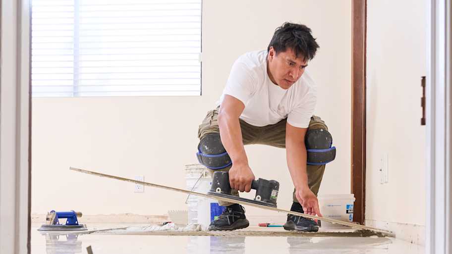A worker in knee pads carefully aligns a tile during floor installation, with a polished floor reflecting his focused posture.