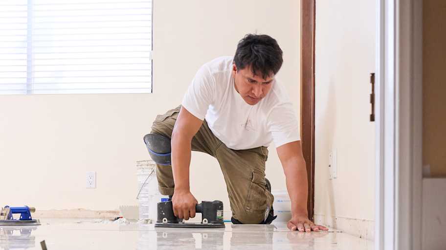 A worker in knee pads aligns a tile on the floor during installation, with his reflection visible on the polished surface.
