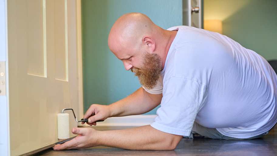 Handyman Chris McNeill from C.A. Shamrock LLC  uses a small roller to carefully paint the bottom edge of a door while lying on the floor for precision.