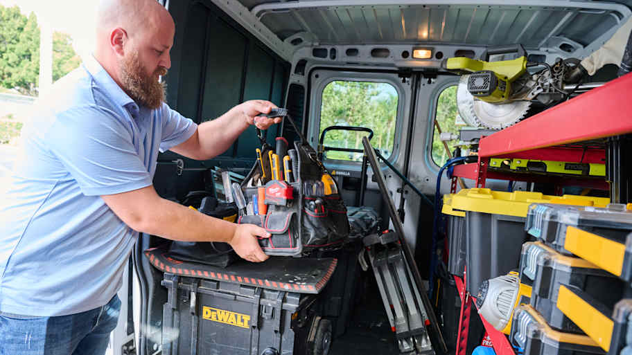 Handyman Chris McNeill from C.A. Shamrock LLC  organizing his tool bag in the back of a work van filled with equipment, toolboxes, and various tools.