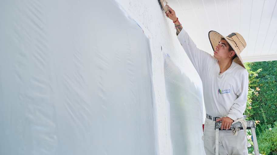 Painter brushes an upper wall corner while standing on a ladder, wearing a straw hat for sun protection.