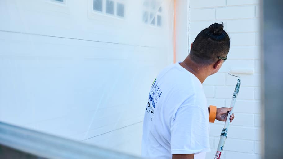 Silvio, a painter with Laguna Painting, uses a roller to paint a brick wall white, with a garage door protected in plastic.