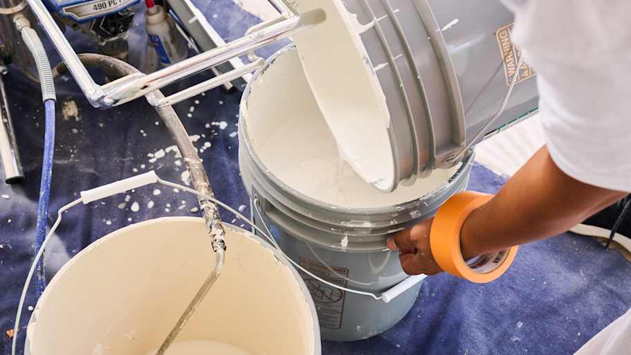 Close-up of a painter pouring white paint into a bucket, preparing supplies for an exterior painting project.