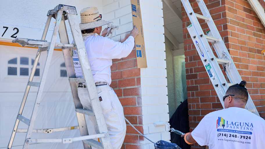 Mario and Silvio from Laguna Painting use ladders to spray paint a brick wall near a garage, carefully protecting surrounding areas.