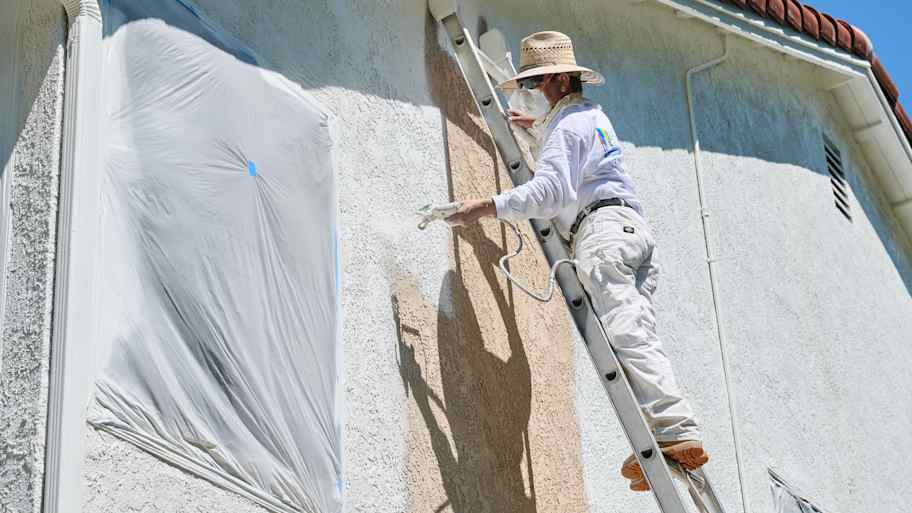 Mario, a painter with Laguna Painting, sprays white paint onto a stucco exterior wall from a ladder, covering the old color.