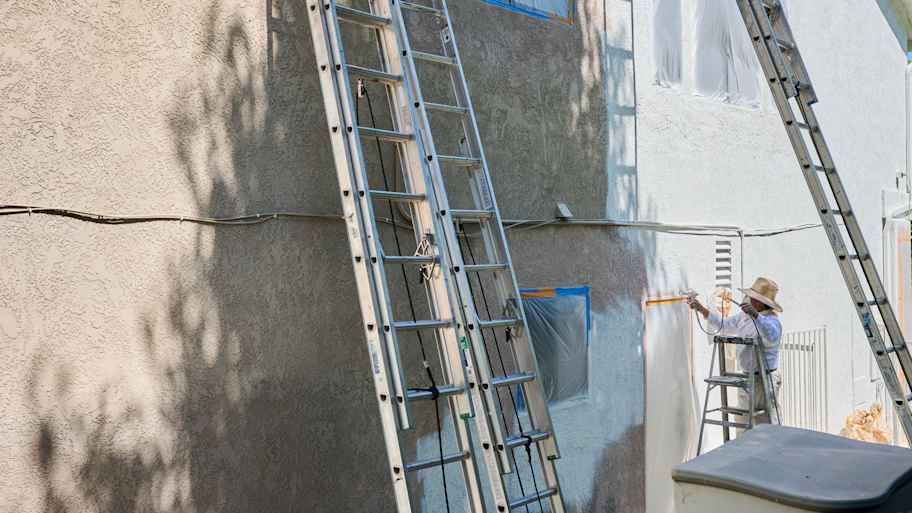 Painter sprays paint on an exterior wall, with ladders and covered windows in view, ensuring an even coat.