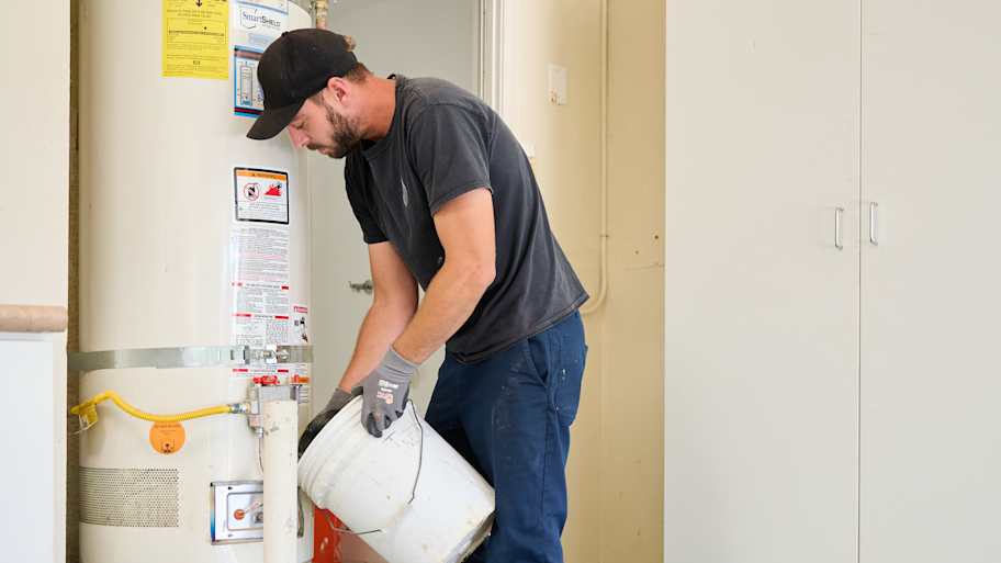 A technician pours water from a bucket near a water heater in a garage, with tools and equipment on the floor around him.