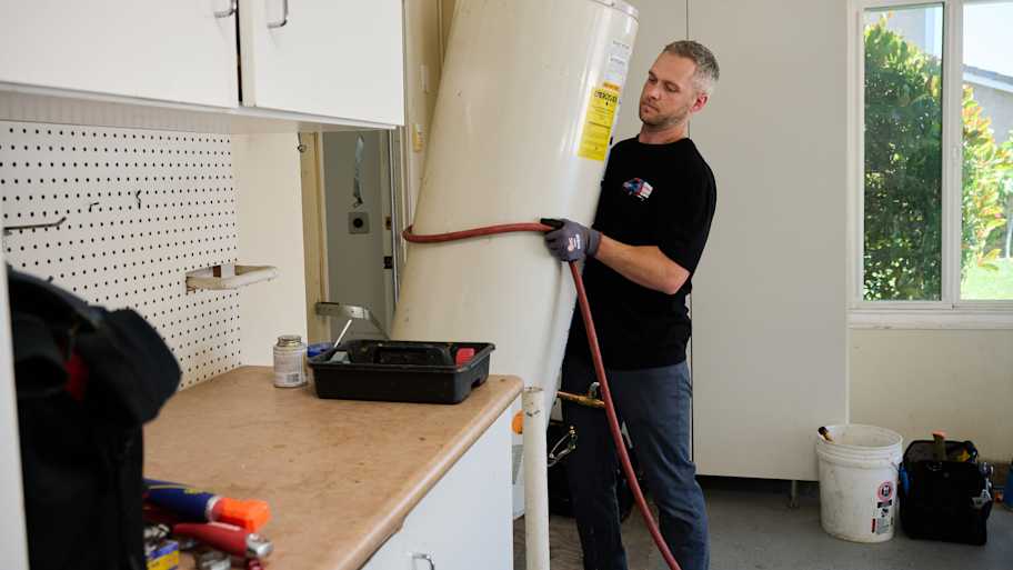 Technician carefully lifts a water heater while wearing gloves in a well-lit garage with tools and supplies on the counter nearby.