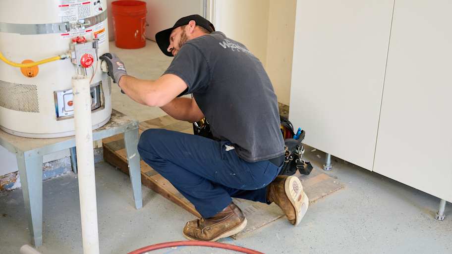 A technician kneels on the floor to inspect a water heater in a garage, surrounded by tools, hoses, and equipment.