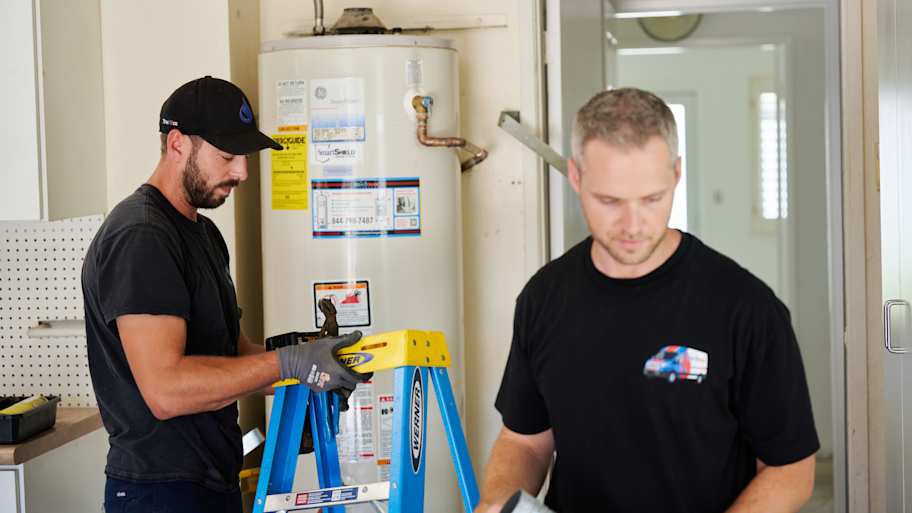 Two technicians work near a water heater; one adjusts tools on a ladder, while the other is focused on equipment in the foreground.
