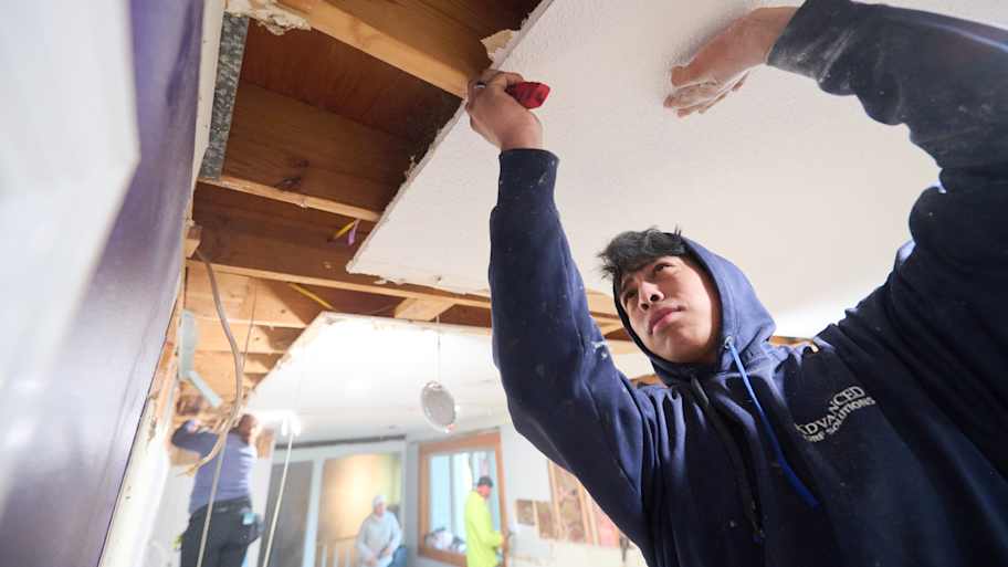 A worker in a blue hoodie carefully removes a ceiling panel to expose wooden beams, with other team members visible in the background.