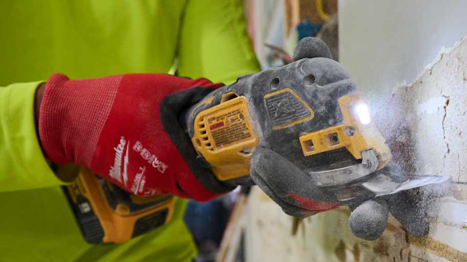 Close-up of a power tool cutting through drywall, with red-gloved hands holding the tool as dust and debris scatter.