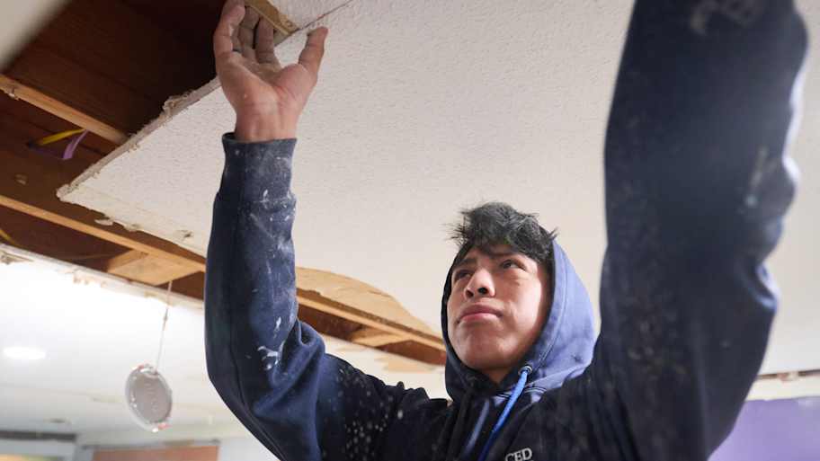 A worker in a blue hoodie carefully removing a section of the ceiling during a renovation project, exposing wooden beams above.