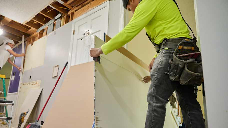 A construction worker measuring drywall while another adjusts its position during a home renovation project.