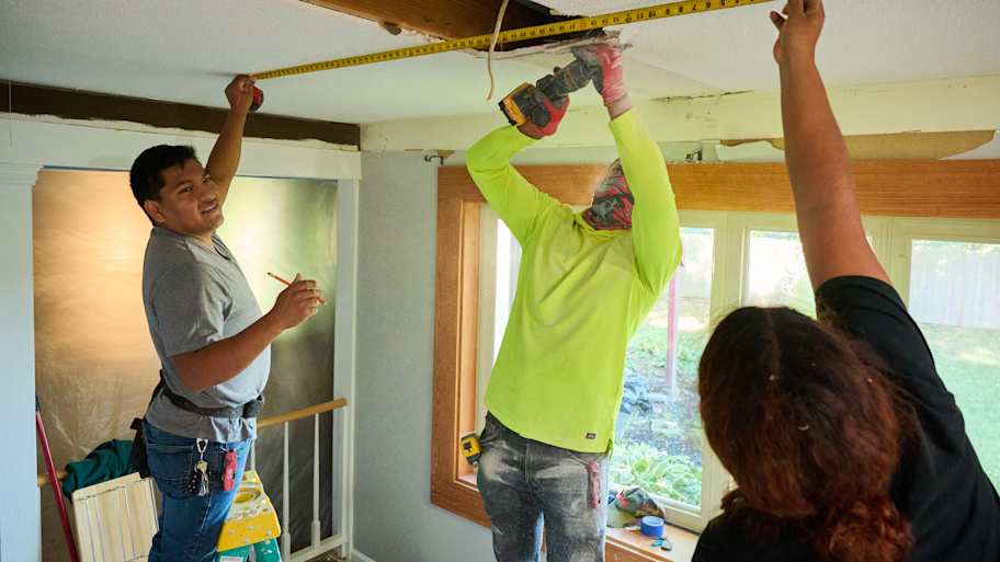 Three workers collaborate on ceiling installation, one measuring with a tape, another drilling, and a third assisting with alignment.