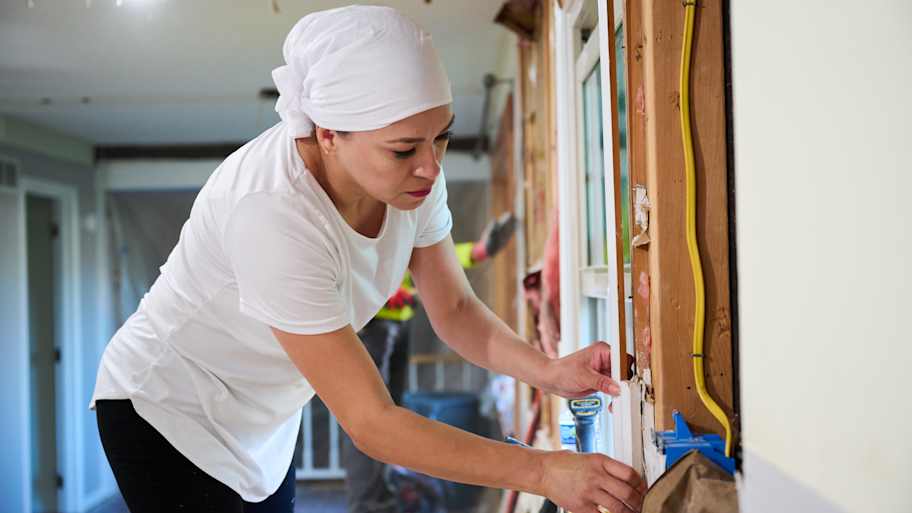 A woman in a white shirt and headscarf carefully works on a window frame during a home renovation, surrounded by exposed walls.