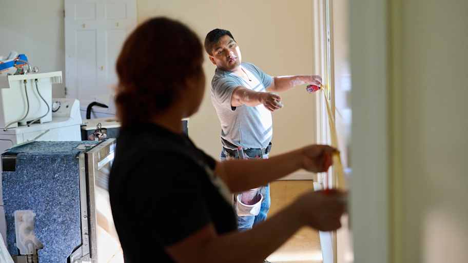 Two workers measuring a wall with a tape measure in a bright room, discussing placement for a renovation project.