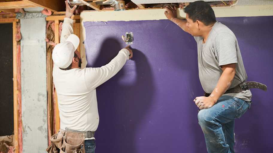 Two workers installing electrical components on a purple wall during a home renovation, with exposed beams and insulation visible.
