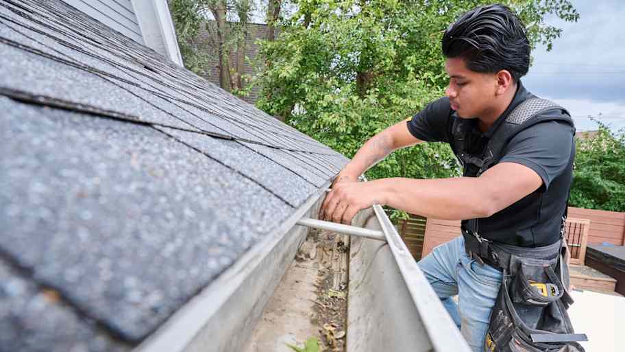 Worker cleaning a clogged roof gutter using a scoop tool, wearing a safety harness, with asphalt shingles overhead.