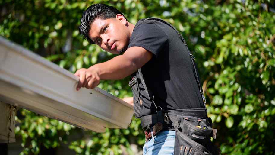 "Worker in a harness examines a white gutter under bright sunlight, surrounded by lush green trees in the background. 