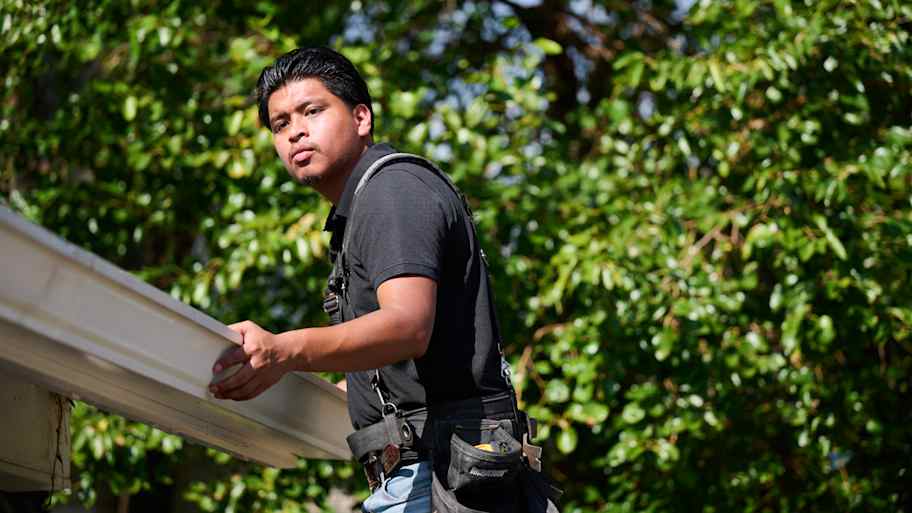 Worker in a tool harness inspects a gutter under bright sunlight, surrounded by green foliage in the background.